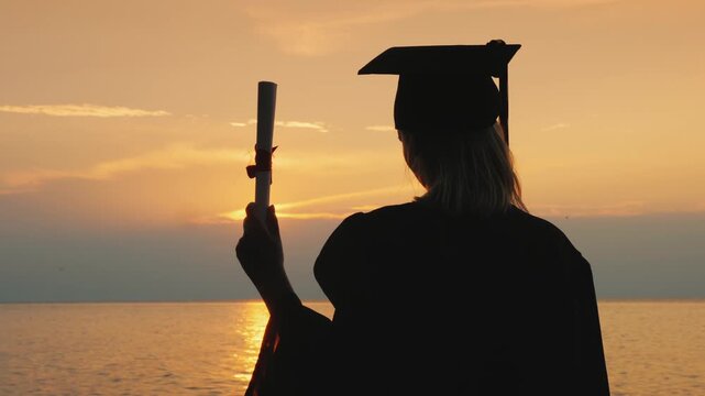 Graduate holding diploma in cap and gown against sunset ocean backdrop celebrating academic achievement.