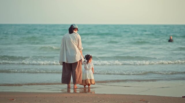 Back view of an Asian mother holding her toddler daughter's hand while standing on the sandy beach, looking at sea waves during sunset. Family bonding, parenting lifestyle, summer travel vacation.