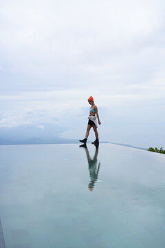 Pensive young woman walking at infinity pool with cloudy sky, Bali