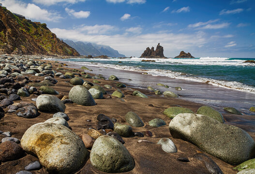 Dramatic shoreline with rounded stones, dark sand, sea waves, and rugged volcanic cliffs beneath, Benijo Beach in the Anaga Rural Park,  Tenerife, Canary Islands.
