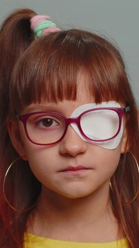 Sick ill girl looking at camera having protective bandage plaster patch scratch on one eye. Injury trauma blind. Child kid wearing glasses at gray background. Medical eyesight vision therapy. Vertical