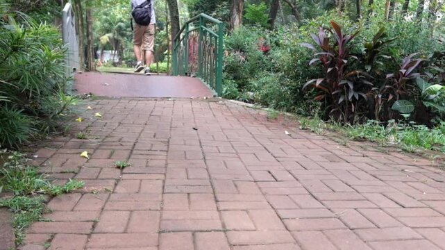 Young man wearing a backpack walks along a paved path in a lush green park setting while moving toward a small metal bridge in this smooth cinematic 4k video clip