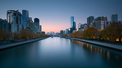 Fototapeta premium River reflects city lights at twilight. Buildings glow softly against dusky sky. Trees line banks, their leaves turning gold. Water flows calmly, mirroring urban grandeur