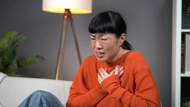 Asian woman suffering from a sudden heart attack, severe chest pain, or panic attack, clutching her chest with both hands. She is on a white sofa at home, visibly distressed with eyes tightly closed.