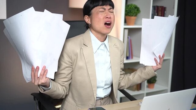Extremely stressed Asian business woman in beige blazer screaming and throwing papers at office desk. Overwhelmed professional lady expressing anger and frustration with workload in workplace.