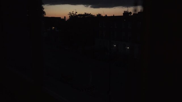 Footage captured from indoors through a window, showing a deep red sunset sky above urban surroundings in London, England. The scene features city silhouettes, warm evening colors, and atmospheric lig