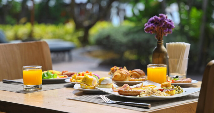 Gourmet breakfast spread sitting on wooden table in outdoor terrace with lush tropical garden background, eggs benedict, croissants, fresh orange juice, bright morning sunlight, luxury resort dining,