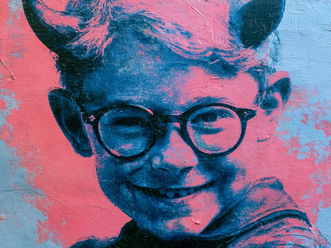 Boy with glasses smiling and wearing devil horns in front of a pink and blue background during the day