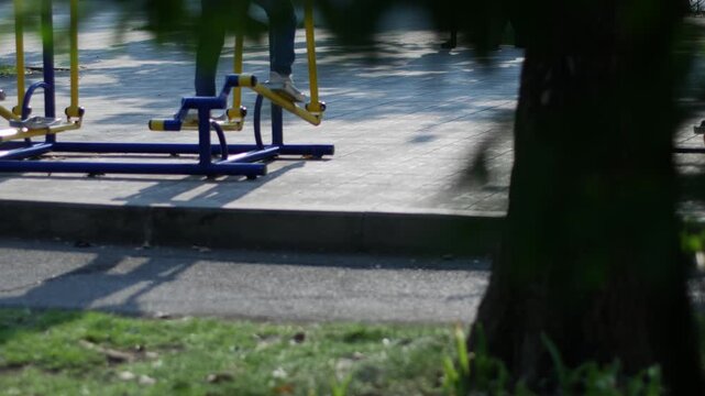 Person using blue and yellow outdoor elliptical exercise machine in a public park with green foliage background and dappled light