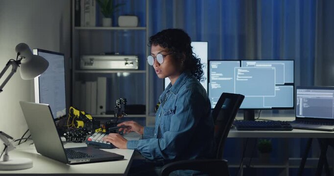 Young African American female programmer coding AI software on multiple screens in night office. Network security, cybersecurity, machine learning, working late, overtime, robotics automation concept.