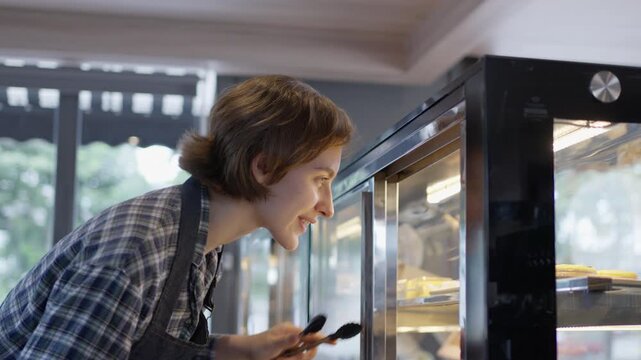 Smiling young woman baker arranging pastry bakery display case, looking food glass display cabinet, apron, cafe interior, focused expression, natural light, small business, SEO friendly video title