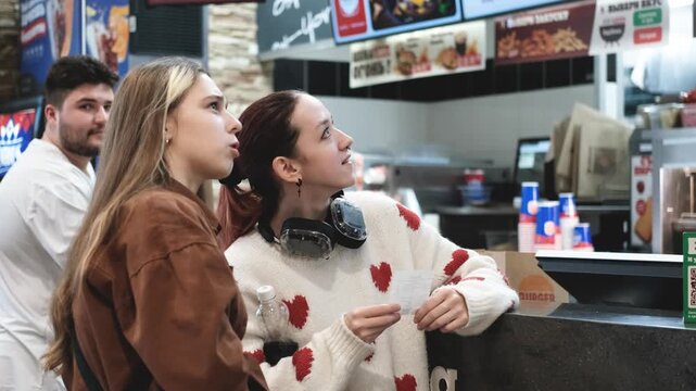Two young women choose food from a menu on a self-service kiosk screen in a fast food restaurant.