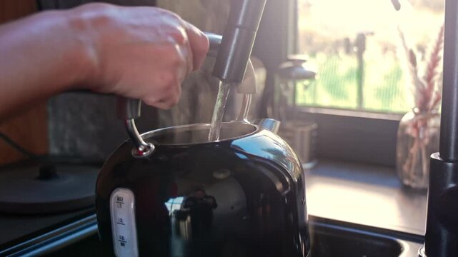 Hand Filling Black Electric Kettle With Water In Kitchen, close up home video frame with steam, sunlight and visible capacity markings