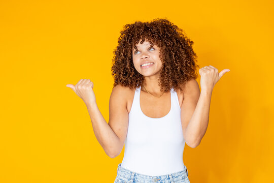 Happy young woman with curly hair pointing both thumbs sideways on bright yellow background, smiling with a fun and playful expression