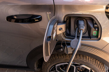 Close-up of an EV charging cable plugged into the charging port of a grey electric car, showing the open fuel flap, tire, and wheel rim. Green energy and modern eco-friendly transport concept. © Micha