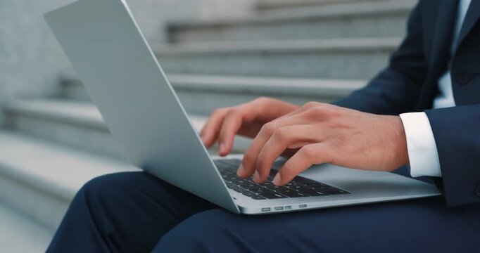 Close up of man in dark blue suit typing on silver laptop while sitting on steps.