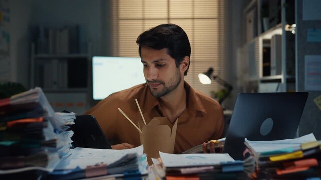 Young Latin businessman taking urgent phone call while reading accounting document on laptop at desk in dark night office. Multitasking overtime, overworked deadline, tired paperwork rush concept.