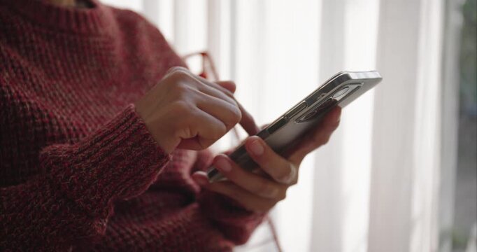Hand. Asian woman using smartphone on chair by window, scrolling screen with finger in bright home interior,