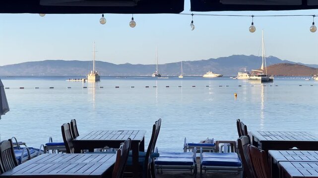 An empty beachfront restaurant terrace in Bitez, Bodrum, looking out over a calm bay filled with anchored yachts. Hanging light bulbs and wooden tables frame a tranquil morning seascape.