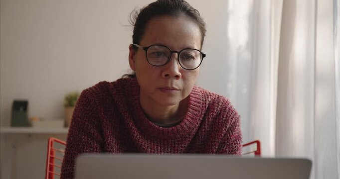 Asian middle woman wear glasses typing work on computer in living room, Front view