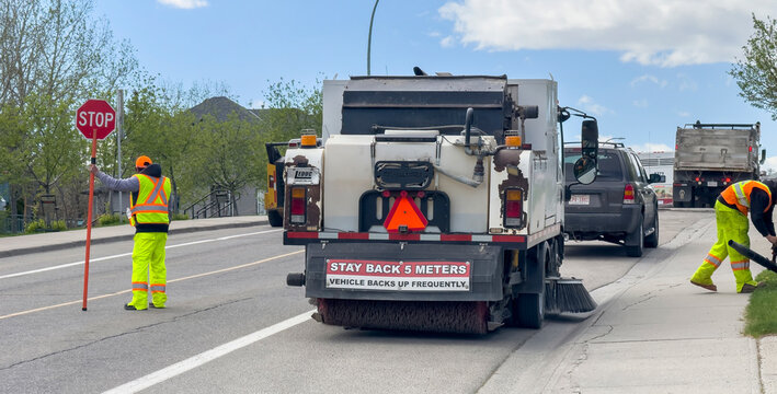 Calgary, Alberta, Canada. May 17, 2026. Municipal street sweeper crew with flagger and blower clearing residential road on sunny spring day.