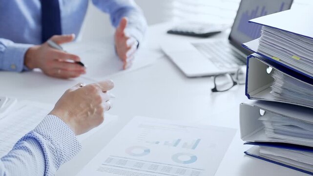 Office workers or auditors are analyzing financial reports and checking documents while sitting at white table with a stack of blue folders in the foreground. Audit and taxes theme in business
