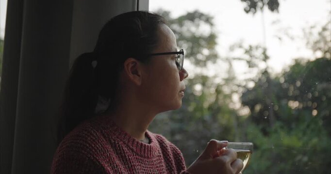 Thoughtful Asian Woman with glasses holding teacup by Window in Morning,  Side View of Relaxed Female Practicing Self-Care and Mindfulness in Cozy Living Room, Slow Living Routine