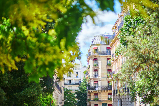 Elegant Paris apartment building with classic balconies and red awnings framed by lush green trees in bright summer light.