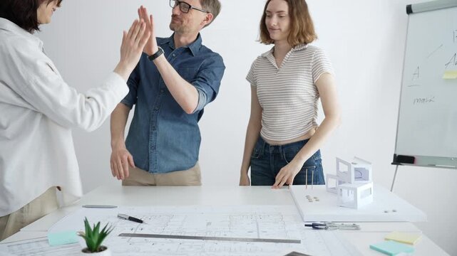 Architecture team collaborating on a project, joining hands as a symbol of unity, teamwork, and success, working on blueprints and a building model in the studio