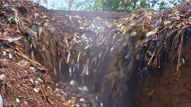 View from inside deep compost trench as soil, roots and organic material collapse downward during outdoor waste disposal work with falling debris, dust clouds and earth movement.