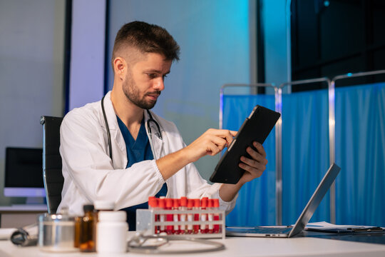 Doctor scientist looking through microscope and holding blood sample tube at workplace.
