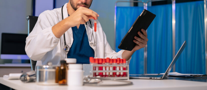 Doctor scientist looking through microscope and holding blood sample tube at workplace.