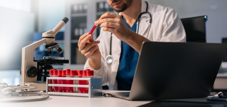 Doctor scientist looking through microscope and holding blood sample tube at workplace.