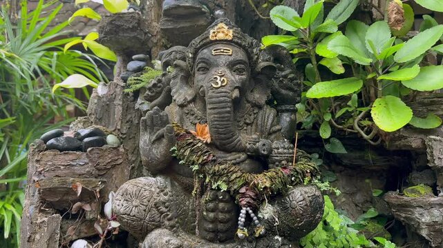 Stone relief of Ganesha decorated with offerings stands among green plants in a traditional religious setting. Traditional worship.