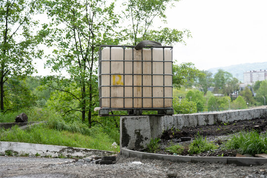 A weathered plastic intermediate bulk container (IBC tote) inside a protective steel grid cage, placed on a concrete wall for water storage or garden watering.
