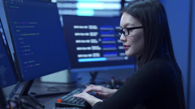 Young female software engineer writing program code on multiple desktop screens in a modern office.