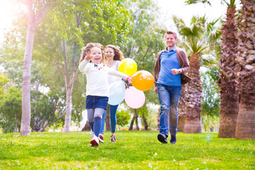 Fototapeta premium A woman and two young girls are running together in a grassy park, smiling and holding colorful balloons on a sunny day. 