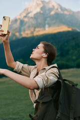 Fototapeta premium Smiling woman with backpack taking a selfie outdoors in front of a dramatic mountain landscape. Casual hiking scene, travel lifestyle, and outdoor exploration with soft natural light.