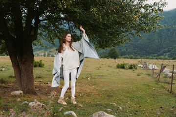 Fototapeta premium A young woman dances in a breezy meadow, wearing white pants and a beige knit sweater with a gray shawl flowing, mountains in the background