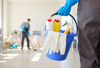Cleaners team office cleaning service with bucket. Blue container holds supplies and gloves as a coworker vacuums. Great for janitorial ads and hygiene content. Clean team service concept now. © Studio Romantic