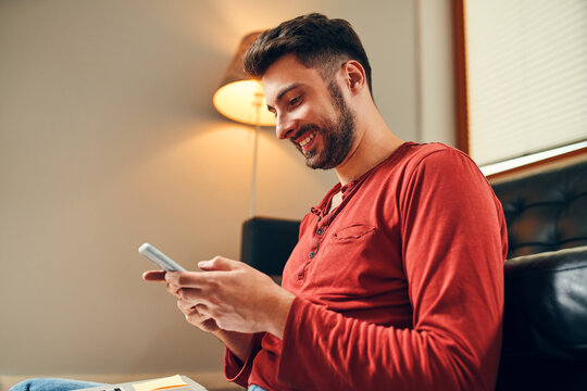 Young male student smiling at smartphone while studying at home