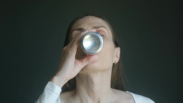Close up portrait thirsty young woman drinking fizzy soda or beer from silver aluminum can, looking directly at camera with satisfied expression after quenching her thirst
