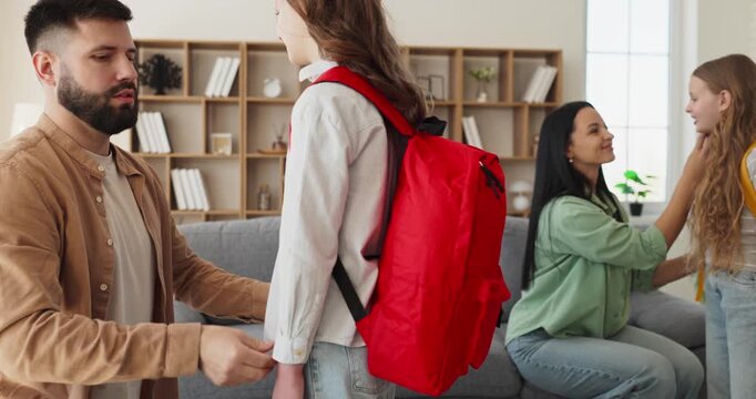 Family parents girls school morning home routine. Father adjusts straps and mother comforts children as backpacks are checked in a cozy living room. Clear back to school preparation.