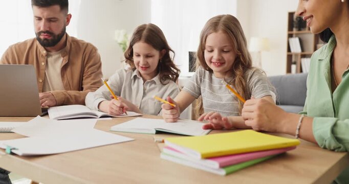 Family parents support children homework studying education. Two girls write in notebooks with pencils as mother helps while father uses a laptop nearby. Concept home learning and support.