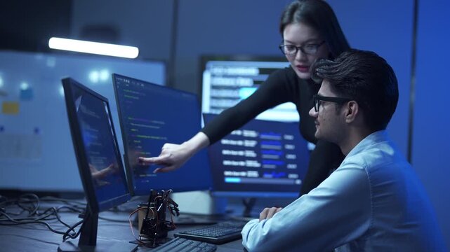Two diverse software programmers working together on a computer program code and digital data metrics on multiple monitors in a modern office.