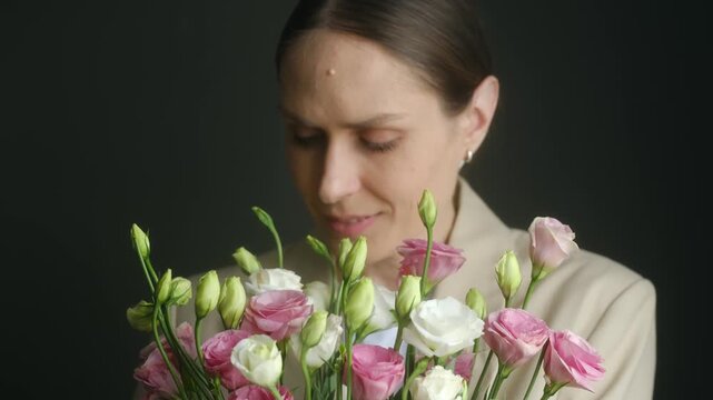 Charming middle aged woman in a business suit smelling a bouquet of fresh pink and white eustoma flowers, smiling with pleasure and happiness against a dark background in a studio