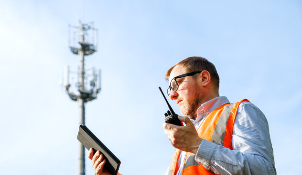 Telecommunications engineer wearing an orange safety vest using a digital tablet and talking on a walkie-talkie. Cell phone tower with 5G antennas stands in the blurred background. Mobile network.