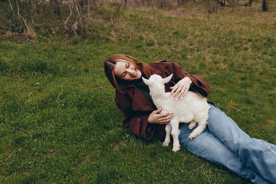 Smiling woman resting on grass, cuddling a young goat kid in a field. Outdoor lifestyle moment showing bond with animals, nature, and wellness. Soft daylight creates a warm, peaceful mood.