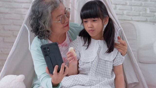 Intimate Bond: A touching moment of connection as a grandmother and grandchild share a heartwarming interaction, the little girl enjoying a snack while looking at a cellphone.