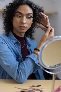 Non-binary adult in denim shirt applying mascara wand while fixing hair at vanity with round mirror